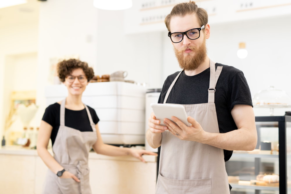 two coworkers in aprons standing in a bakery using a tablet