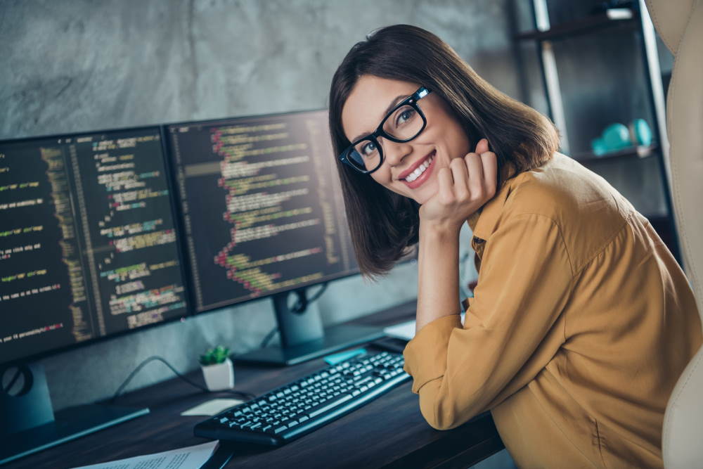 Frau mit Brillen arbeitet vor einem Computer mit mehreren Bildschirme auf denen ein HTML Code zu sehen ist und lacht in die Kamera
