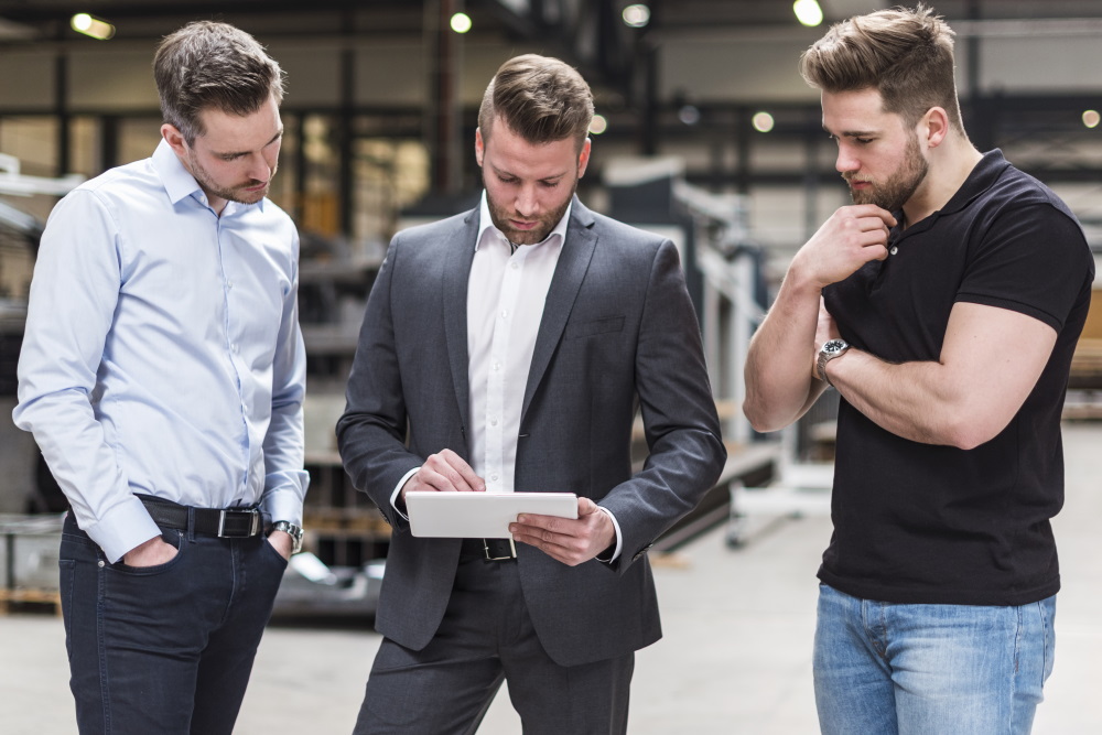 Drei Männer stehen in einer Werkstatt und schauen auf ein Tablet